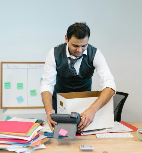 A man packing up his desk in an office setting with papers and a phone.